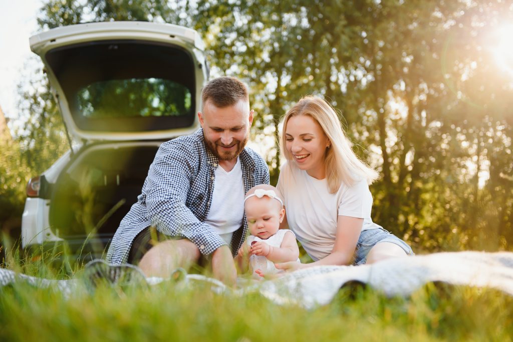 mladá rodina s malým dieťatkom na pikniku v prírode, v pozadí otvorené auto